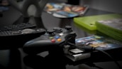 A gaming controller with colorful buttons is placed on a reflective surface, alongside a remote control. Gaming discs and cases are visible in the background, giving a sense of a gaming setup.