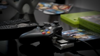 A gaming controller with colorful buttons is placed on a reflective surface, alongside a remote control. Gaming discs and cases are visible in the background, giving a sense of a gaming setup.