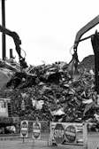 Two large industrial machines with claws are moving a pile of metal scraps and debris. In the foreground, several signs say 'STOP' and 'DO NOT ENTER'. The machinery is surrounded by heaps of twisted metal and broken materials.