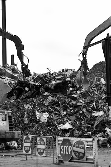 Two large industrial machines with claws are moving a pile of metal scraps and debris. In the foreground, several signs say 'STOP' and 'DO NOT ENTER'. The machinery is surrounded by heaps of twisted metal and broken materials.