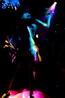 A dynamic and colorful scene featuring a silhouetted figure dancing under vibrant lights. The image captures the movement and energy through streaks of neon pink, blue, and yellow against a dark background.