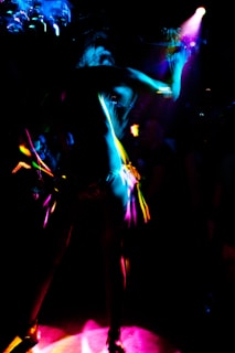 A dynamic and colorful scene featuring a silhouetted figure dancing under vibrant lights. The image captures the movement and energy through streaks of neon pink, blue, and yellow against a dark background.