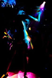 A dynamic and colorful scene featuring a silhouetted figure dancing under vibrant lights. The image captures the movement and energy through streaks of neon pink, blue, and yellow against a dark background.