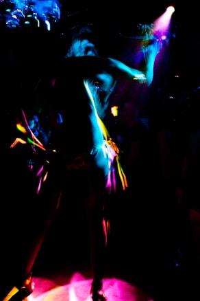 A dynamic and colorful scene featuring a silhouetted figure dancing under vibrant lights. The image captures the movement and energy through streaks of neon pink, blue, and yellow against a dark background.