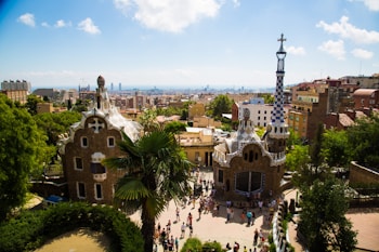 A vibrant cityscape with unique architecture featuring whimsical, mosaic-covered structures nestled among lush greenery. Numerous people are seen walking along the winding paths, enjoying the sunlit surroundings. In the distance, a sprawling urban landscape extends towards the horizon, with the sea barely visible under a bright sky.