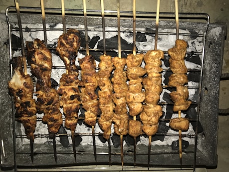 Several skewers of grilled meat are arranged over a bed of charcoal on a barbecue grill. The meat appears to be well-cooked with a slightly charred exterior.