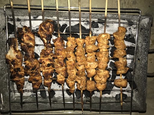 Several skewers of grilled meat are arranged over a bed of charcoal on a barbecue grill. The meat appears to be well-cooked with a slightly charred exterior.