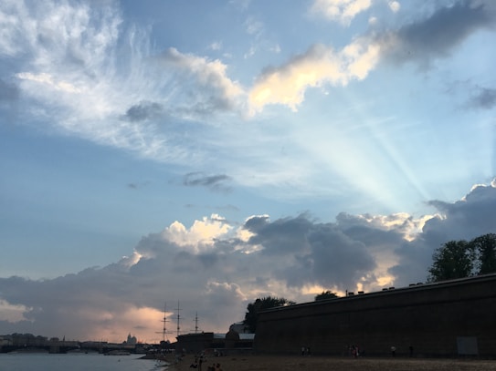 The image features a vast sky filled with dramatic clouds, some letting through rays of sunlight, creating a picturesque and serene atmosphere. Below the sky, there's a waterfront scene with a beach and a distant view of ships with masts silhouetted against the horizon. A fortress wall stands to the right, giving a historical feel.