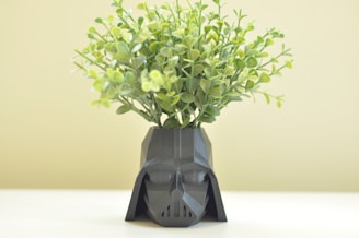 A geometric, dark-colored planter shaped like a helmet with leafy green plants sprouting from the top. The background is a soft, light color.