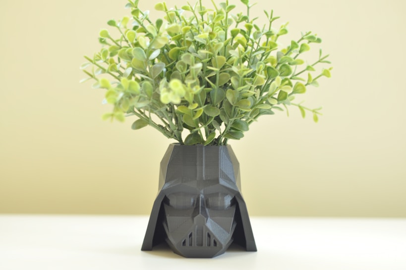 A geometric, dark-colored planter shaped like a helmet with leafy green plants sprouting from the top. The background is a soft, light color.