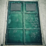 A large, weathered green metal door with rust and peeling paint is set in a textured concrete wall. Two rectangular ventilation panels with a grid pattern are located at the top of each panel of the door, suggesting an industrial or utilitarian use.
