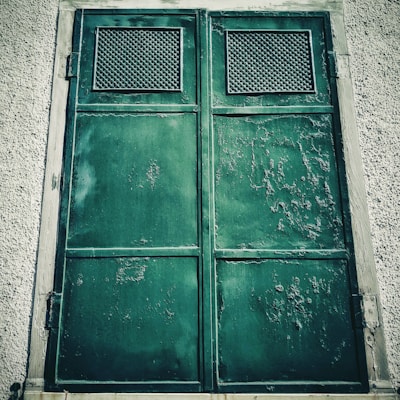 A large, weathered green metal door with rust and peeling paint is set in a textured concrete wall. Two rectangular ventilation panels with a grid pattern are located at the top of each panel of the door, suggesting an industrial or utilitarian use.