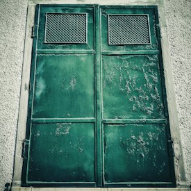 A large, weathered green metal door with rust and peeling paint is set in a textured concrete wall. Two rectangular ventilation panels with a grid pattern are located at the top of each panel of the door, suggesting an industrial or utilitarian use.