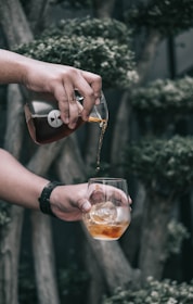 A person is pouring an amber-colored liquid from a glass carafe into a clear glass with ice cubes. The background features blurred greenery and trees, suggesting an outdoor setting.