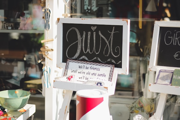 A charming display featuring small chalkboards with artistic cursive writing and a framed sign with a sentimental quote. Colorful decorative objects, such as a green bowl and patterned items, are arranged around the display. The setup is likely part of a storefront or shop window with a playful, inviting vibe.