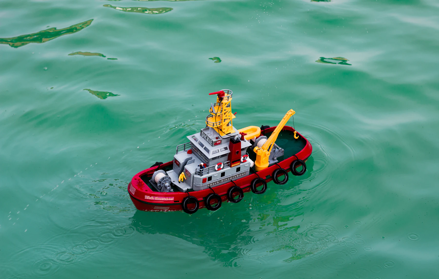 A red and gray model fireboat tug with tire fenders under way on green water
