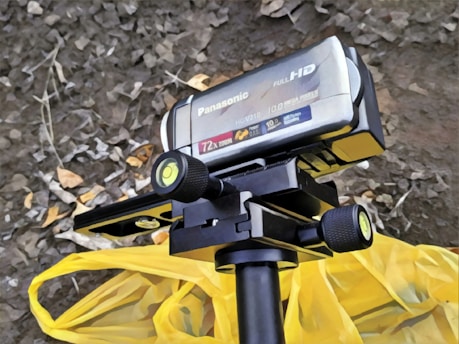 A black Panasonic Full HD camcorder is mounted on a tripod. It has various features labeled on it, including 72x zoom and 10.0 Mega Pixels. The camcorder is positioned above a yellow fabric or bag on a ground covered with dry leaves.