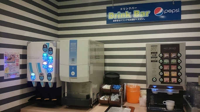 Two beverage dispensers are against a wall with black and white stripes. The left dispenser has illuminated blue buttons, likely for cold drinks. To the right, another machine offers hot drink options with labeled buttons. Beneath the machines, various supplies such as cups, lids, and stirrers are organized, with an orange container holding trash. A sign above reads 'Drink Bar' with a Pepsi logo.