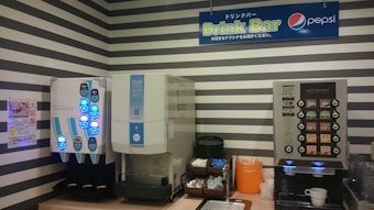 Two beverage dispensers are against a wall with black and white stripes. The left dispenser has illuminated blue buttons, likely for cold drinks. To the right, another machine offers hot drink options with labeled buttons. Beneath the machines, various supplies such as cups, lids, and stirrers are organized, with an orange container holding trash. A sign above reads 'Drink Bar' with a Pepsi logo.