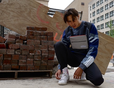A person crouches near a stack of bricks on the street, wearing a blue and white striped sweater, dark pants, and sneakers. They have a gray chest bag labeled 'UTOPIA' and are adjusting their shoelaces. Behind them, there is a wooden board with some graffiti, and buildings are visible in the background.