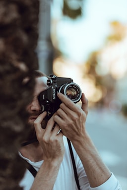 A person is holding a camera up to their face, seemingly focused on capturing a photograph. The surroundings are slightly blurred, with natural elements suggesting an outdoor setting. The person's concentration is evident as they adjust the camera settings.