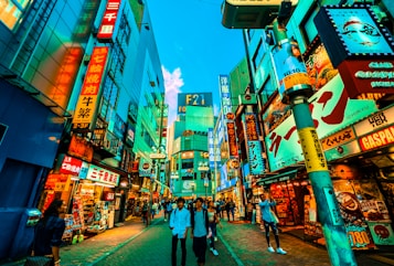 Vibrant city street bustling with people walking past an array of colorful, neon-lit storefronts and billboards. The scene is lively with a mix of modern architecture and bright signage featuring Japanese characters. The sky is a vivid blue, adding contrast to the rich hues of the signs and buildings.