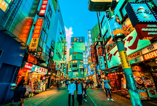 Vibrant city street bustling with people walking past an array of colorful, neon-lit storefronts and billboards. The scene is lively with a mix of modern architecture and bright signage featuring Japanese characters. The sky is a vivid blue, adding contrast to the rich hues of the signs and buildings.