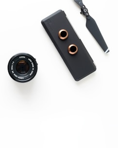 An arrangement of photography equipment including a Canon camera lens, a matte black camera accessory with copper-colored rings, and a drone propeller, all set against a white background.