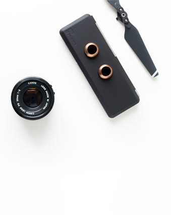 An arrangement of photography equipment including a Canon camera lens, a matte black camera accessory with copper-colored rings, and a drone propeller, all set against a white background.
