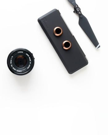 An arrangement of photography equipment including a Canon camera lens, a matte black camera accessory with copper-colored rings, and a drone propeller, all set against a white background.