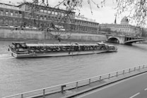 A long, glass-roofed riverboat cruises along a river with a stone embankment and large historic building in the background. Bare tree branches hang down from above, partially framing the scene. An arched bridge is visible in the distance, connecting the two sides of the river.