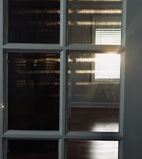 Sunlight streams through blinds, reflecting on a set of multi-paneled glass windows. The light casts distinct horizontal shadows that create a warm pattern against dark glass and wooden flooring.