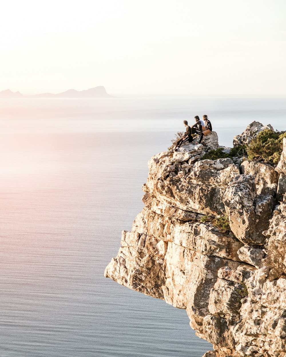 three persons sitting on gray concrete rock mountain under gray sky