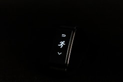 A digital fitness tracker with a black band is displayed against a dark background. The screen features minimalistic white icons for navigation and an exercise mode, including an arrow, a running figure, and a downward-pointing arrow.