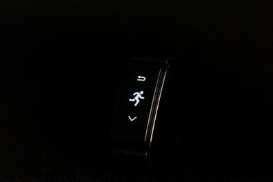 A digital fitness tracker with a black band is displayed against a dark background. The screen features minimalistic white icons for navigation and an exercise mode, including an arrow, a running figure, and a downward-pointing arrow.