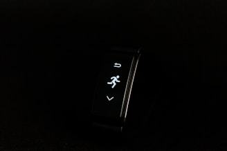 A digital fitness tracker with a black band is displayed against a dark background. The screen features minimalistic white icons for navigation and an exercise mode, including an arrow, a running figure, and a downward-pointing arrow.