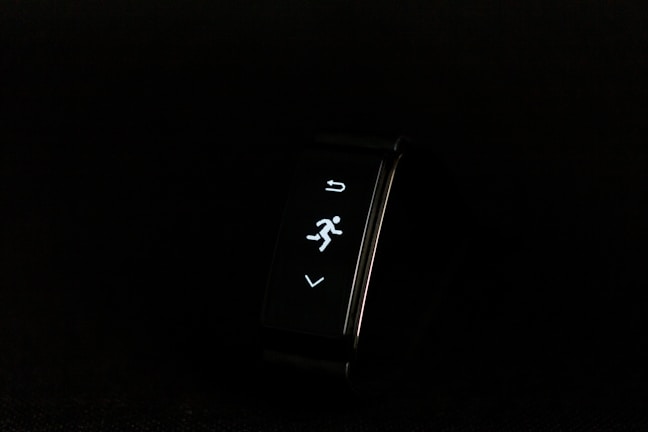 A digital fitness tracker with a black band is displayed against a dark background. The screen features minimalistic white icons for navigation and an exercise mode, including an arrow, a running figure, and a downward-pointing arrow.