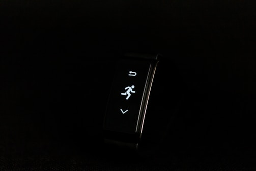 A digital fitness tracker with a black band is displayed against a dark background. The screen features minimalistic white icons for navigation and an exercise mode, including an arrow, a running figure, and a downward-pointing arrow.