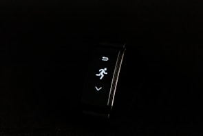 A digital fitness tracker with a black band is displayed against a dark background. The screen features minimalistic white icons for navigation and an exercise mode, including an arrow, a running figure, and a downward-pointing arrow.