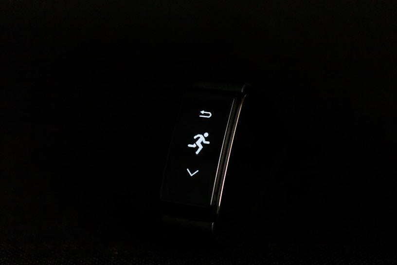 A digital fitness tracker with a black band is displayed against a dark background. The screen features minimalistic white icons for navigation and an exercise mode, including an arrow, a running figure, and a downward-pointing arrow.