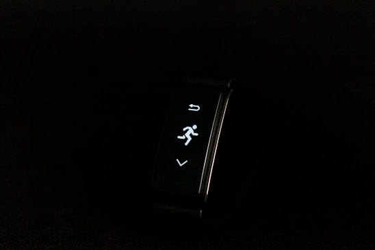 A digital fitness tracker with a black band is displayed against a dark background. The screen features minimalistic white icons for navigation and an exercise mode, including an arrow, a running figure, and a downward-pointing arrow.