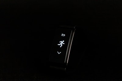 A digital fitness tracker with a black band is displayed against a dark background. The screen features minimalistic white icons for navigation and an exercise mode, including an arrow, a running figure, and a downward-pointing arrow.