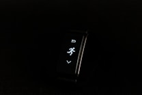 A digital fitness tracker with a black band is displayed against a dark background. The screen features minimalistic white icons for navigation and an exercise mode, including an arrow, a running figure, and a downward-pointing arrow.