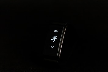 A digital fitness tracker with a black band is displayed against a dark background. The screen features minimalistic white icons for navigation and an exercise mode, including an arrow, a running figure, and a downward-pointing arrow.