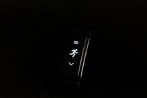 A digital fitness tracker with a black band is displayed against a dark background. The screen features minimalistic white icons for navigation and an exercise mode, including an arrow, a running figure, and a downward-pointing arrow.