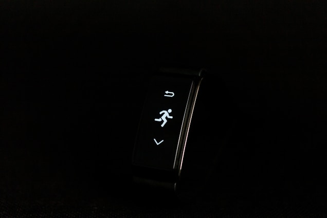 A digital fitness tracker with a black band is displayed against a dark background. The screen features minimalistic white icons for navigation and an exercise mode, including an arrow, a running figure, and a downward-pointing arrow.