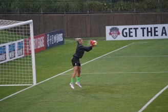 A soccer goalkeeper in motion as they catch a ball mid-air during a game, wearing a black jersey and neon green socks. The scene is set on a well-maintained grass field, and it appears to be raining lightly. Advertising boards and a goalpost frame the background.