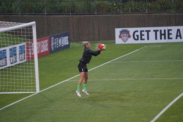 A soccer goalkeeper in motion as they catch a ball mid-air during a game, wearing a black jersey and neon green socks. The scene is set on a well-maintained grass field, and it appears to be raining lightly. Advertising boards and a goalpost frame the background.