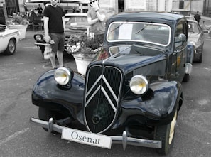 A vintage car with a shiny, polished surface reflecting the surroundings. The car is parked in an outdoor setting, possibly a car show, with other cars and people in the background. The surroundings include potted plants and a building with visible windows in the background.