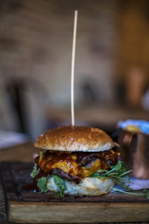 A juicy cheeseburger topped with melted cheddar cheese, barbecue sauce, and fresh greens, placed on a wooden board. A tall skewer pierces through the center, and a blurred background suggests a rustic setting.